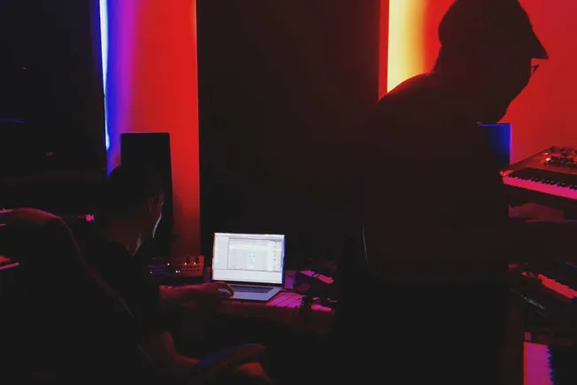 Creative music production studio with musicians working on digital audio workstations and synthesizers in a dimly lit, colorf