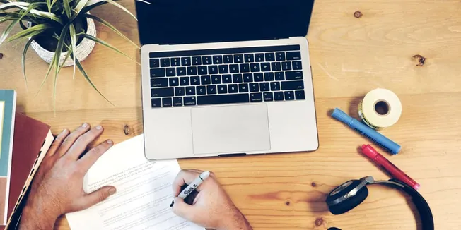 High-angle view of a workspace with a laptop, open book, and writing tools on a wooden desk, emphasizing digital music indust