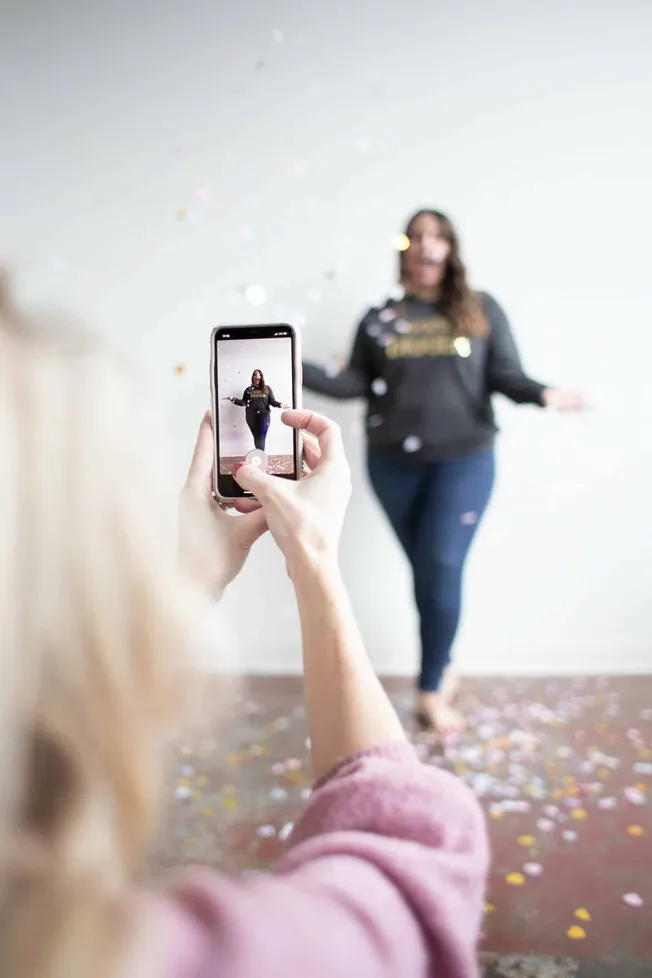 Teen woman taking a photo of friend with smartphone at celebration, fun party atmosphere, confetti, social media, capturing m