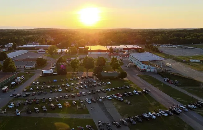 Sunset over outdoor concert venue with parked cars, stage, and surrounding buildings in a rural area, capturing live music ev