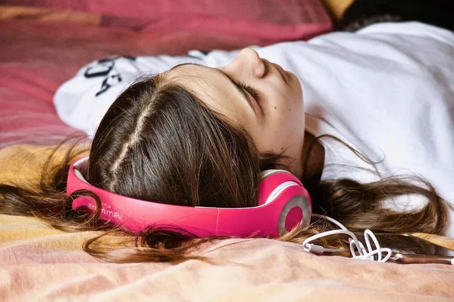 Pink over-ear headphones resting on a young woman's head as she lies on a bed with her eyes closed, relaxing and listening to