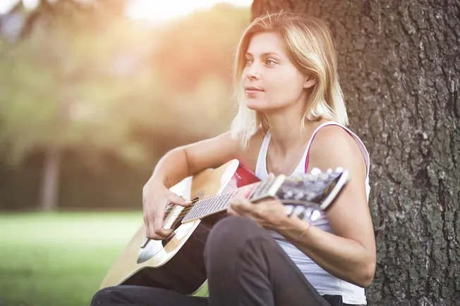 Girl playing acoustic guitar outdoors in park during sunset, relaxing music session, young woman with blonde hair, casual clo
