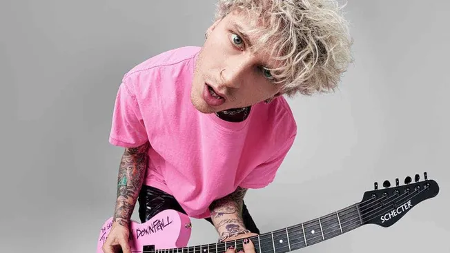 Viral musician with platinum blonde curly hair wearing a pink t-shirt playing a pink Schecter guitar against a plain gray bac