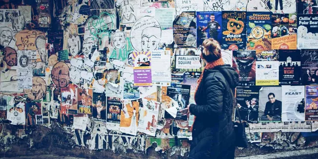 Poster wall full of various colorful flyers and advertisements with a woman in a black coat looking at the posters in an urba