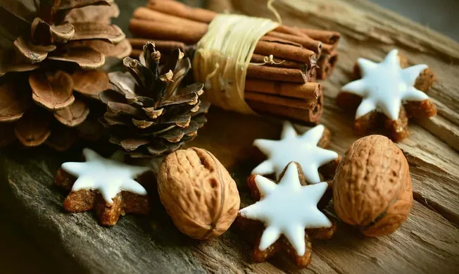 Sprigs of cinnamon, star-shaped cookies with white icing, walnuts, and pinecones arranged on rustic wooden surface for holida