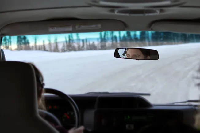 Frosty winter road viewed from inside a vehicle with a driver and a woman looking through the rearview mirror, snowy landscap