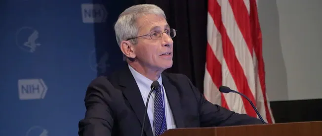 Senior male speaker at a press conference or panel discussion, with an American flag and NH logo in the background, deliverin