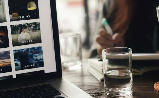 Photo of a laptop displaying social media images, glass of water, and person taking notes at a wooden table in a cozy cafe en