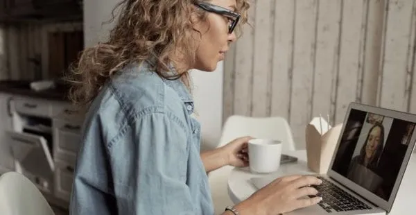 A woman participating in a virtual meeting on her laptop, with a coffee mug nearby, in a cozy workspace setting.