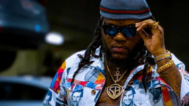 Rapper with dreadlocks wearing sunglasses and colorful jacket featuring celebrity portraits, accessorized with gold chains an