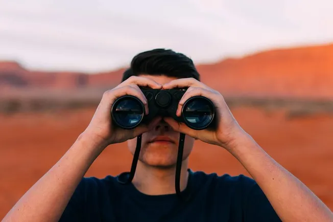 A young man looking through binoculars in a desert landscape with red rock formations in the background.