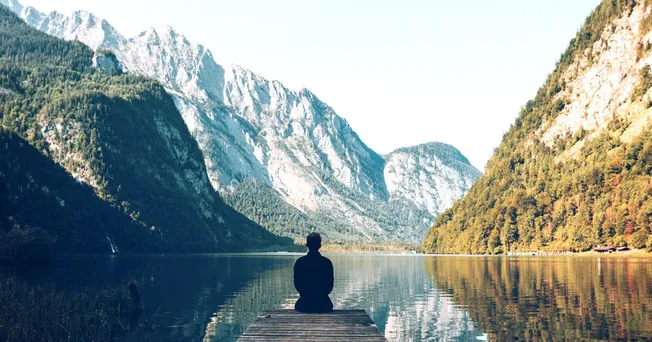 Serene person meditating by mountain lake surrounded by lush green forest and towering peaks, inspiring outdoor adventure, na