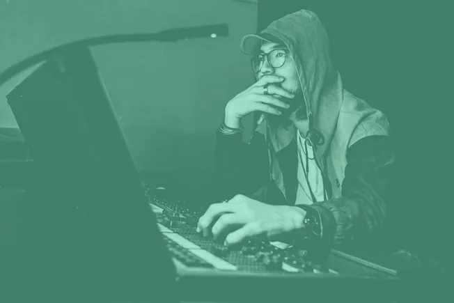 Young man working on music production, DJing, or audio editing at a computer with headphones and a hoodie, emphasizing digita