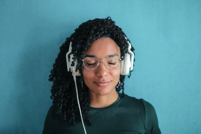 Relaxed woman listening to music with headphones, wearing glasses and a dark shirt, standing against a blue background.