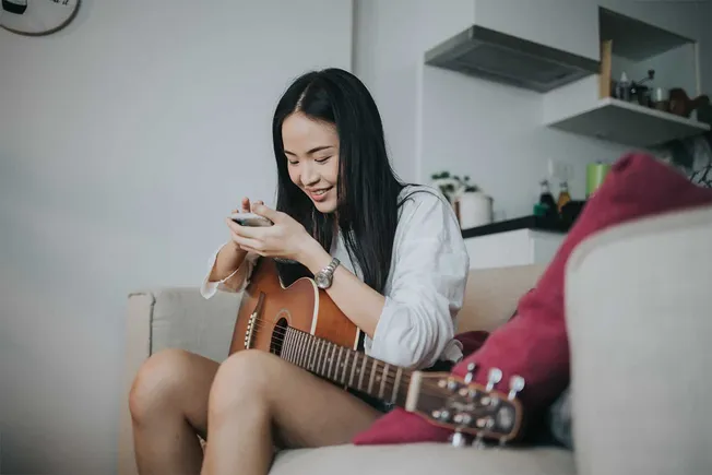 Young woman playing acoustic guitar while using smartphone at home, creative music practice, casual lifestyle, relaxed atmosp