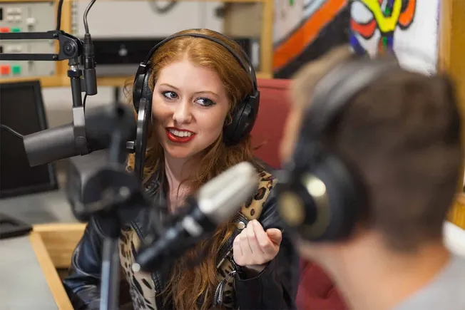 Female radio host speaking into microphone during podcast recording, wearing headphones, in a studio setting with colorful ar