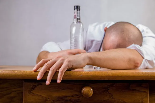 Feeling exhausted or overwhelmed, a man with a bald head is resting his head and arms on a wooden table, with an empty beer b