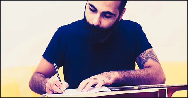 A young man with a beard and tattoos writing in a notebook, sitting on a yellow couch, engaging in creative work or music com