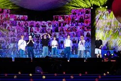 Performers on stage at a music conference or concert, with vibrant lighting and large digital screens displaying visuals behi