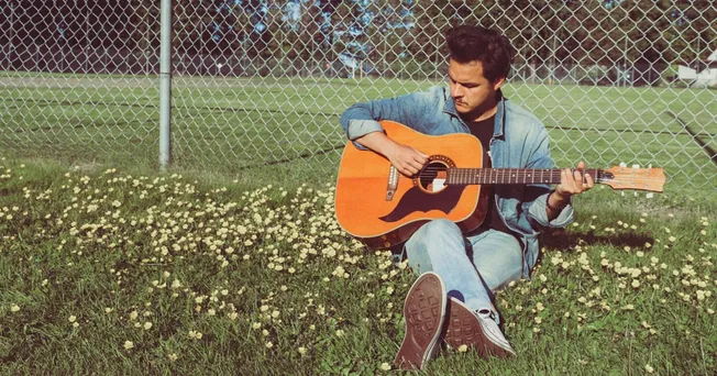 Guitarist sitting on grass near a chain-link fence, playing acoustic guitar outdoors in a park setting, capturing musical cre