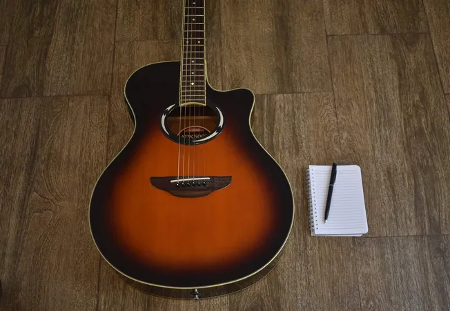 A vintage acoustic guitar with a sunburst finish lying on wooden flooring next to a small spiral notepad and black pen, ideal