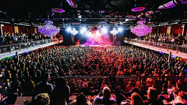 Vibrant concert hall filled with a large crowd, colorful stage lights, and elegant chandeliers, showcasing live music perform
