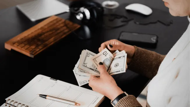 $50, $20, and $10 bills held in person's hands on desk with notebook, pen, headphones, and electronic devices, representing m