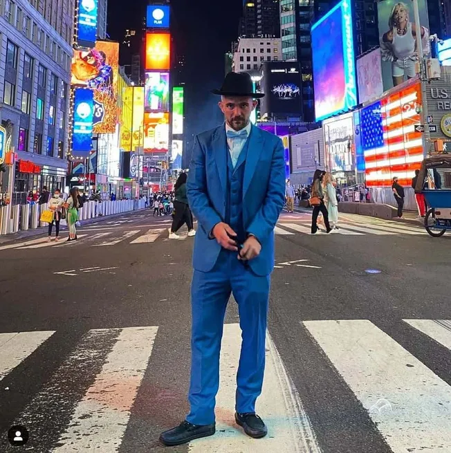 Neo-noir man in blue suit and black fedora standing on crosswalk in Times Square at night, illuminated billboards, vibrant ci