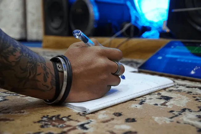 Hand with tattoos writing in notebook on wooden table music and creative workspace environment.