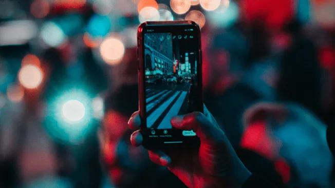 A person capturing a city street scene with a smartphone during nighttime, illuminated by vibrant city lights and bokeh effec