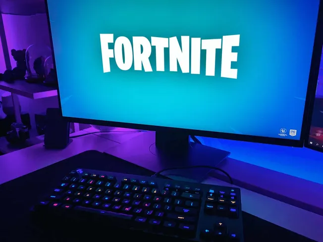Gaming computer setup featuring a large monitor displaying the Fortnite logo, with colorful RGB keyboard and gaming accessori