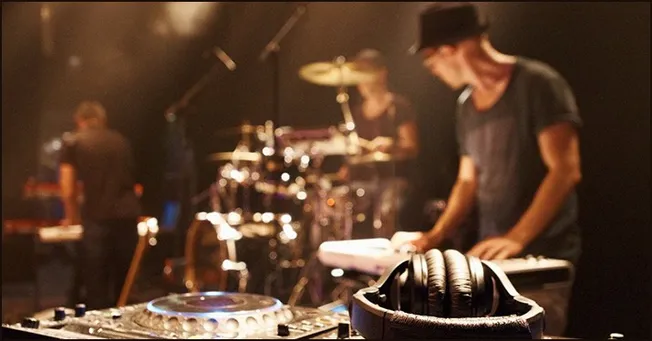 DJ equipment and headphones on stage during a live musical performance by a band with drummer and keyboardist in the backgrou