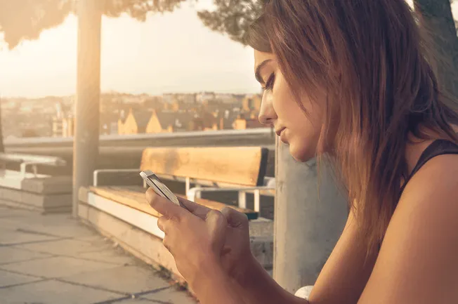 Young woman using smartphone outdoors during sunset, sitting on park bench, enjoying social media or messaging.