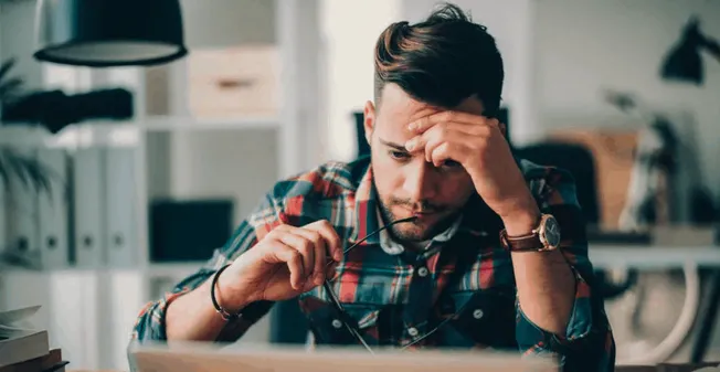 Frustrated young man at desk overwhelmed with work, stress, and digital fatigue, feeling anxious about deadlines and overwhel