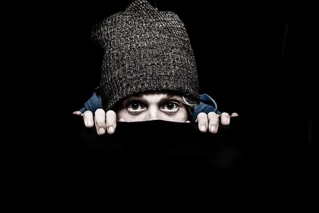 Wary young person peeking from dark background, wearing a knit beanie, emphasizing privacy and curiosity, suitable for digita