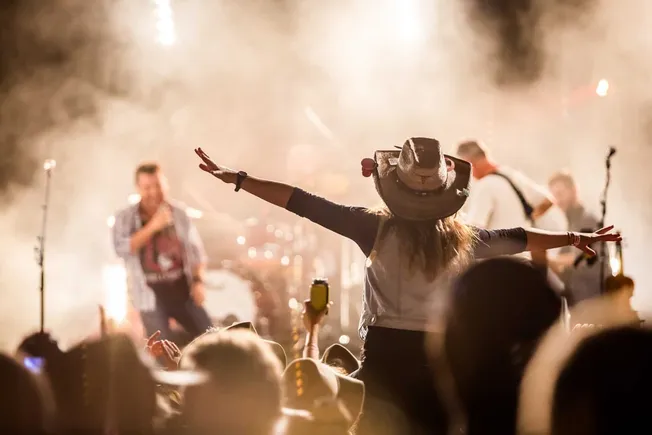 Vibrant concert scene with a female festival attendee wearing a wide-brimmed hat and outstretched arms, enjoying live music a