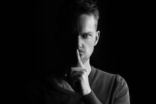 A man with serious expression holding finger to lips in a dark, mysterious setting.