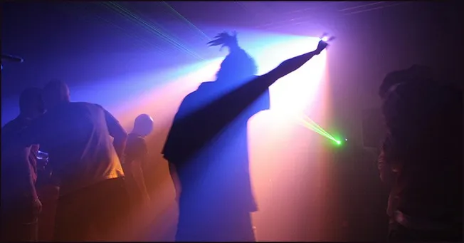 Dance floor party scene with colorful lights and silhouettes of people enjoying a nightclub or concert atmosphere.