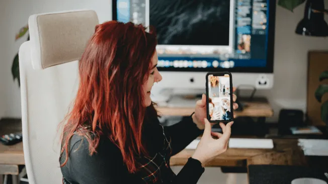 Woman taking a selfie in front of a computer monitor displaying music editing software at a desk with creative workspace setu