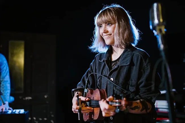 Ukulele player smiling on stage in a dark concert setting, performing live music, young female musician with guitar, music pe