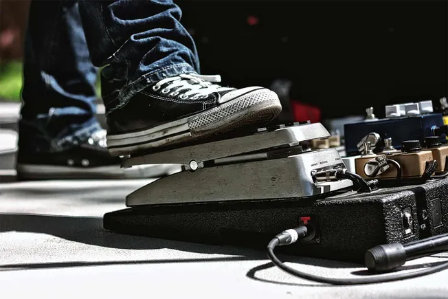 Foot of a musician wearing black Converse sneakers pressing on a guitar effects pedal with multiple pedals in the background,