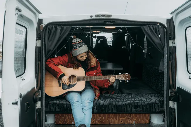 Cozy musician playing acoustic guitar inside a van during winter, woman in red jacket and beanie, urban outdoor scene, chilly