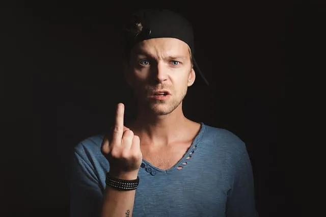 Upset man in baseball cap making rude gesture, expressing anger or frustration, dark background, casual clothing, modern styl