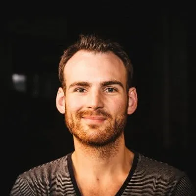 Handsome young man with short brown hair and a beard, smiling confidently in a dark background.