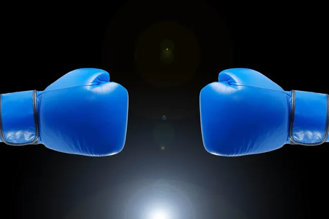 Blue boxing gloves facing each other against a black background, symbolizing competition or conflict, digital image related t