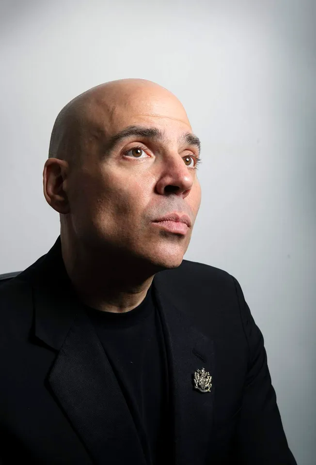 Bald man in a black suit with a police badge looking thoughtfully to the side against a plain background.