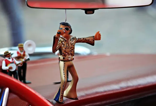 Retro Elvis Presley air freshener hanging from car rearview mirror with mini band figurines in the background.