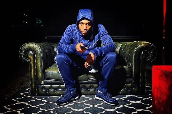 Young man in a blue hoodie and tracksuit sitting on a vintage leather sofa, holding gaming controllers and a phone, in a dark