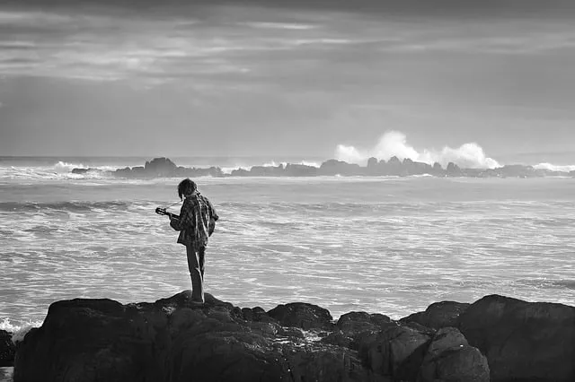 A young person standing on rocks by the ocean, holding a smartphone and looking at the water, captured in black and white.