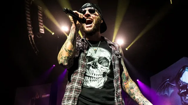 Male musician performing live on stage with tattoos, skull graphic t-shirt, black cap, and sunglasses under colorful concert